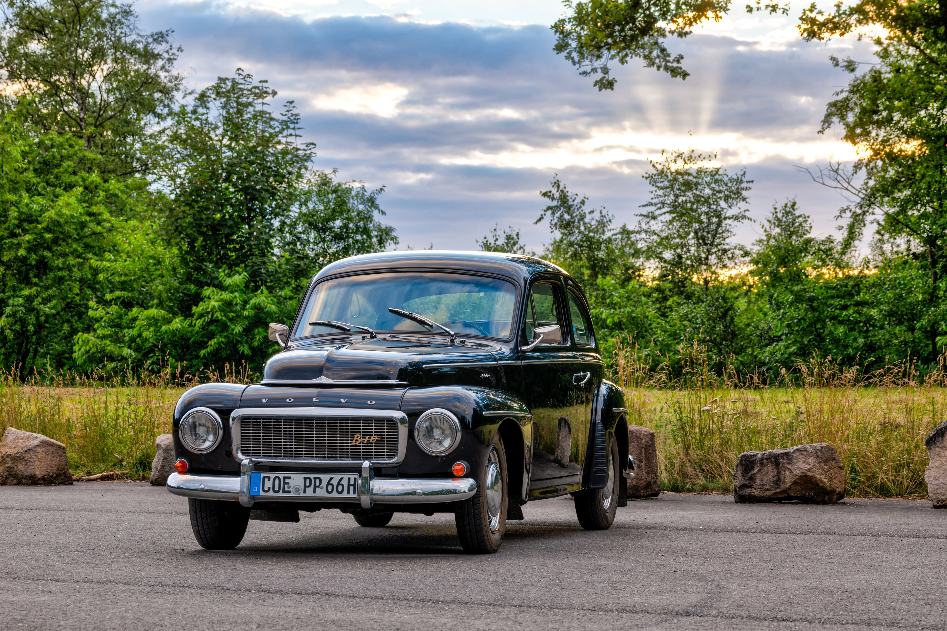 Schwarzer Volvo PV 544 von 1961 auf einem Parkplatz vor Bäumen und Abendhimmel, vorne das H-Kennzeichen COE-PP 66H
