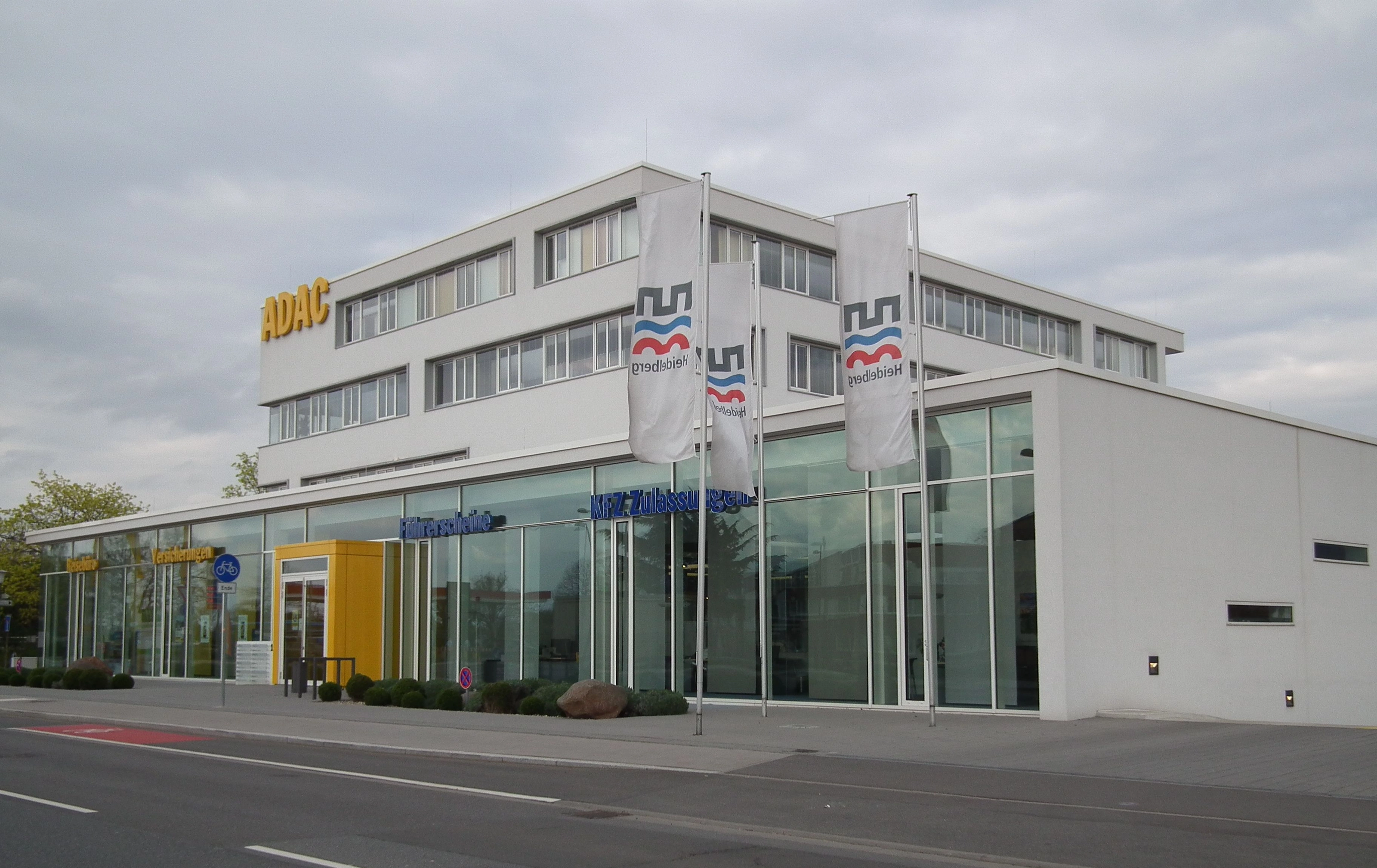 Modernes Bürogebäude mit Glasfront und dem Schriftzug KFZ-Zulassungsstelle, davor Fahnen der Stadt Heidelberg und eine Straße