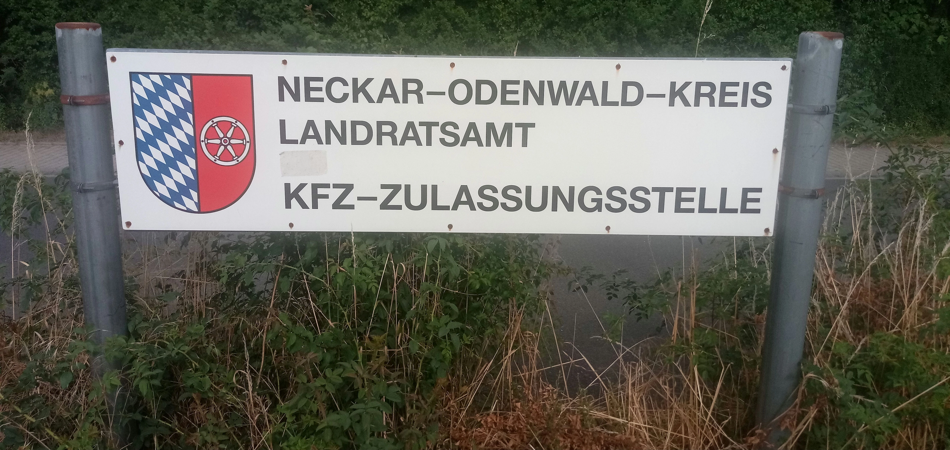 Verwittertes weißes Hinweisschild zwischen zwei Metallpfosten vor Gebüsch mit Kreiswappen und der Aufschrift „Neckar-Odenwald-Kreis Landratsamt Kfz-Zulassungsstelle“