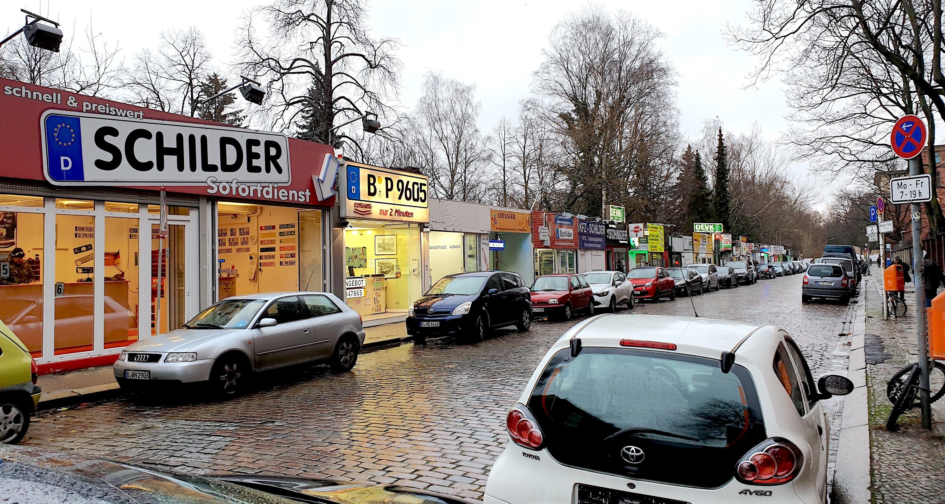 Straße mit einer Reihe von Schilderläden vor der Kfz-Zulassungsstelle in Berlin; über dem vordersten Laden ein großes Schild in Kennzeichenform mit der Aufschrift SCHILDER, davor parkende Autos auf nassem Kopfsteinpflaster