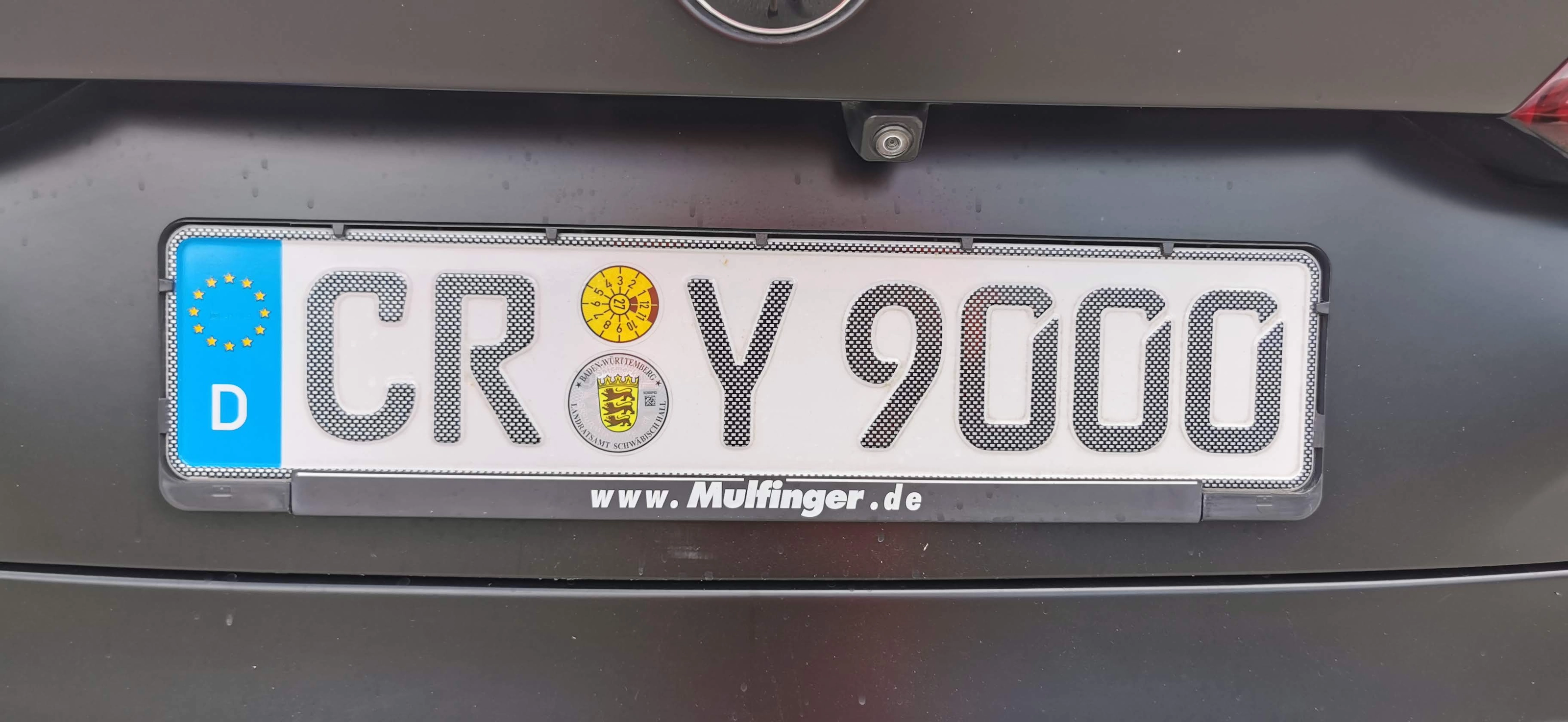 Nahaufnahme eines deutschen Kennzeichens CR Y 9000 am Heck eines dunklen Autos, mit blauem EU-Band und D, gelber HU-Plakette und Stempelplakette des Landratsamts Schwäbisch Hall