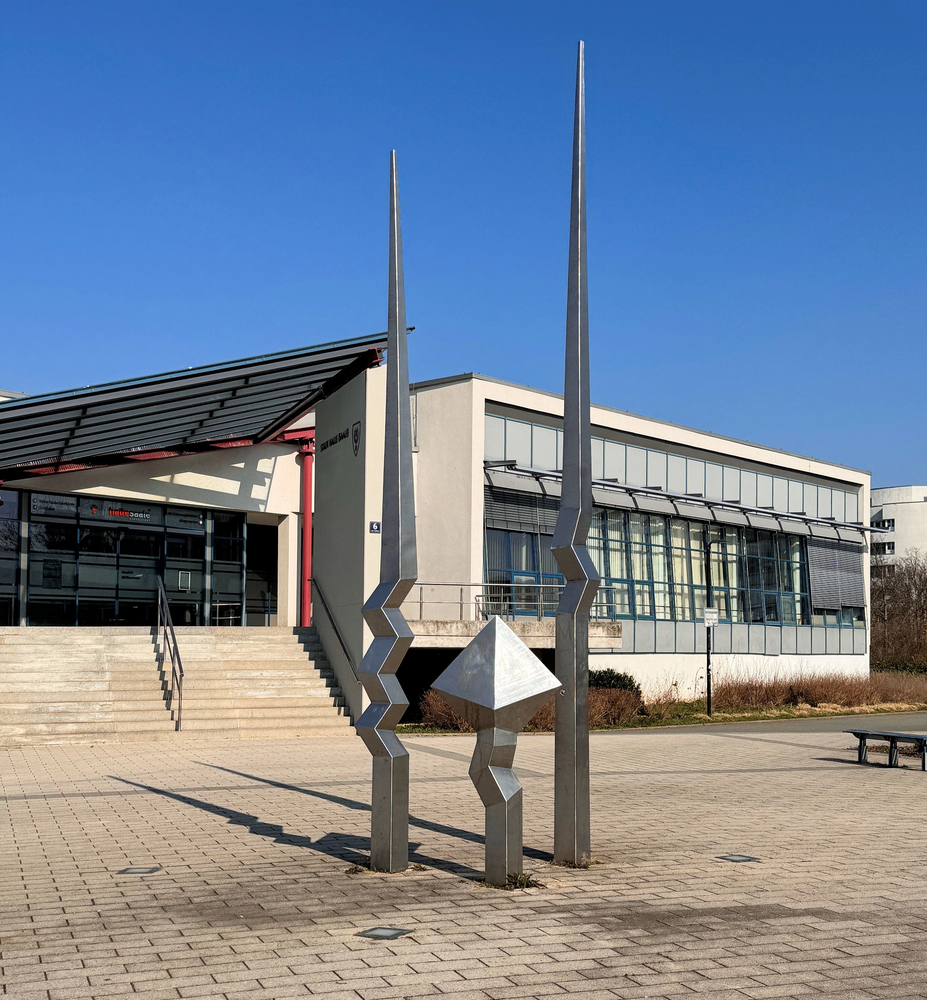 Flaches Verwaltungsgebäude der Kfz-Zulassungsstelle in Halle (Saale) mit Freitreppe und Glasfront, davor auf einem gepflasterten Platz eine Edelstahlskulptur aus drei Stelen unter blauem Himmel